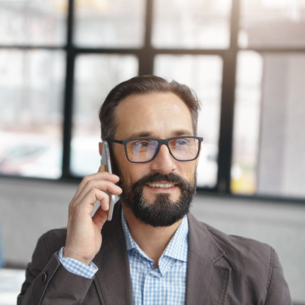 Close up portrait of bearded mature businessman has telephone conversation with partner, discuss future transactions or net capital gain, stands against office interior, people work in background