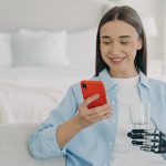Smiling young disabled woman holding smartphone and glass of pure water. Female with bionic prosthetic arm using mobile phone apps, chatting in social networks, shopping online at home.