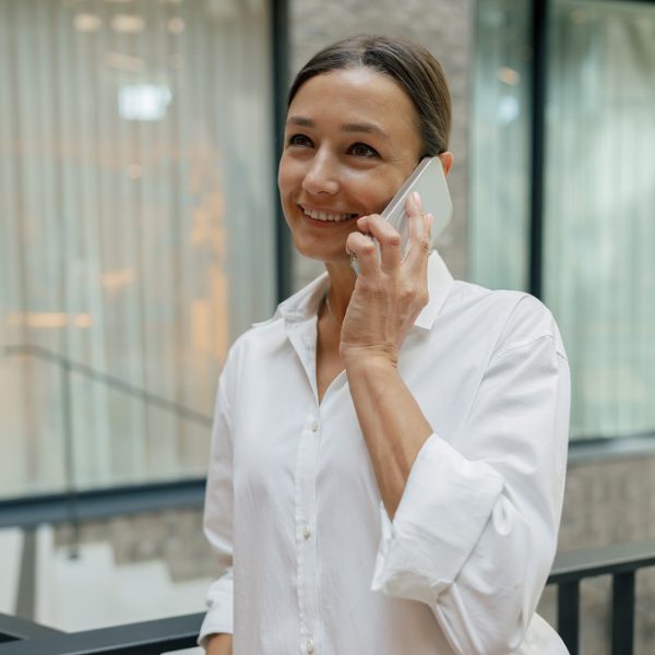 Cheerful woman entrepreneur talking phone with client while standing on modern office background
