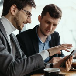 One-on-one meeting. Two young business people sitting at table in restaurant having a conversation using a phone and having a coffee. Businessman using a smartphone